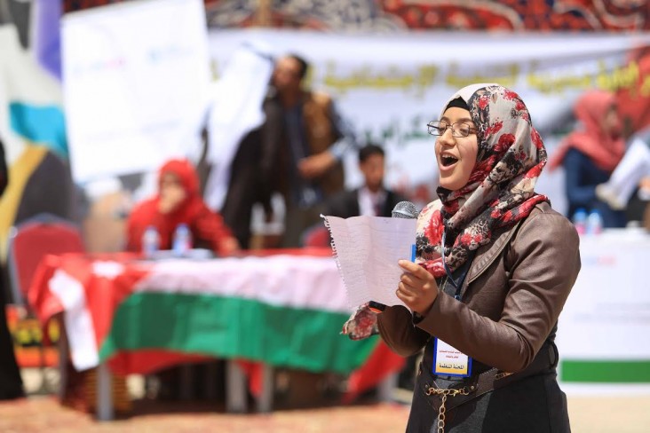 A young women speaks with a microphone in front of a table covered with a Jordanian flag. 