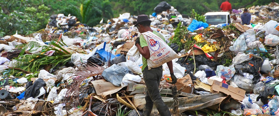 A man walks through a garbage dump—a sack slung over his shoulder in one hand, a bundle of wire in the other—salvaging waste with residual value.